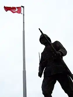 Photographie en contre-plongée d'une statue en bronze représentant un soldat tenant un fusil et un sabre. Un drapeau turc au sommet d'un mat est visible à l'arrière-plan.