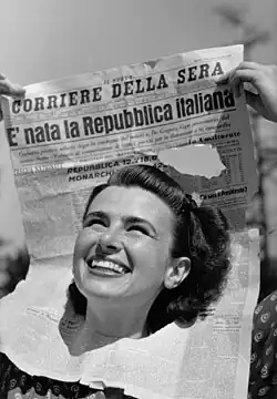 Jeune femme souriante brandissant au-dessus de sa tête le titre d'un exemplaire du Nuovo Corriere della Sera du 6 juin 1946 titrant en italien "La République italienne est née". La femme a enfilé jusqu'au cou le journal par un trou dans la moitié inférieure de ce dernier.
