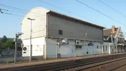 Bâtiment technique de forme parallélipédique dans le prolongement du bâtiment voyageurs de la gare d'Épône - Mézières, avec un soubassement en maçonnerie blanc et un étage sans ouvertures, au toit bombé, recouvert ainsi que l'étage de bardage métallique gris clair, au premier plan 2 voies et caténaires