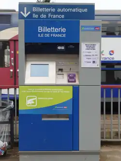 Appareil distributeur automatique de billets de couleurs bleue et grise  portant l'indication « Billetterie automatique Île-de-France ». En arrière-plan, train de banlieue à deux niveaux.