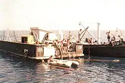 Photographie couleur argentique. En mer un pneumatique au premier plan. Deux barges à couple chargent un morceau de bois encore dans l'eau.