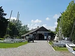 photo de paysage ; au fond, un bâtiment de type entrepôt, auquel mène une route en graviers. de part et d'autres de la route, des bateaux (catamarans et monocoques) sont garés ; un tracteur stationné sur la route est utilisé pour les déplacer