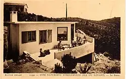 Carte postale noir et blanc. Vue surplombant un bâtiment à flanc de coteau de plein pied avec une terrasse sur laquelle se trouvent des adultes atablés et un enfant dans une voiture à pédales.