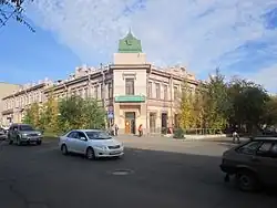 Bâtiment de deux étags situé à l'angle de deux rues. Il est de couleur rose pâle et présente une architecture classique avec des fenêtres rectangulaires, un toit plat et quelques éléments décoratifs. L'entrée du bâtiment à l'angle des deux rues est surmonté d'un petit dôme vert. Des arbres et des arbustes sont plantés autour du bâtiment ainsi que quelques piétons et des voitures circulent sur les rues adjacentes, sur fond de ciel bleu avec quelques nuages.