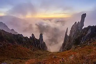 Des rochers pointus et découpés se lancent vers le ciel au milieu d'une mer de nuage. Un sol presque à pic est couvert d'une végétation rase, ocre et verte.