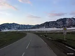 Route dans une vallée avec un virage, avec en arrière-plan des montagnes. Les hauteurs sont enneigées, mais la vallée ne l'est qu'en partie.