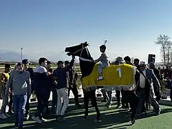 De la foule entoure un cheval de course noir monté par un enfant.