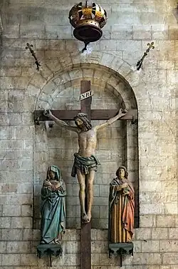 Photo couleur d'un Christ en croix encadré de femmes en prière. La croix est fixée dans une alcôve du mur à linteau roman.