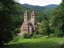 Photographie d'un paysage de montagnes recouvertes de forêts avec, au centre, un bâtiment en pierre entouré par des arbres et composé de deux hautes tours carrées ayant chacune un toit triangulaire