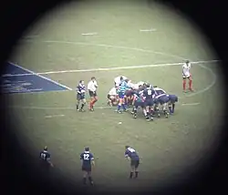Prise de vue lointaine avec un cercle entouré de noir pour se focaliser sur une mêlée du match France - Écosse. Les avants sont en position, les demis en mouvement autour. La phase de jeu n'est pas encore entamée.