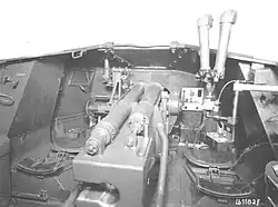 photographie en noir et blanc montrant l’intérieur d’un char sans toit, avec la culasse du canon à l’avant-plan et un périscope proéminent à droite