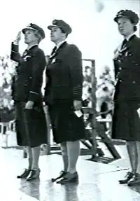 Trois femmes en uniforme militaire sombre, debout au garde-à-vous sur une estrade.