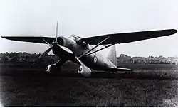 Photo en noir et blanc d'un avion à hélice posé sur une surface en herbe, à l'arrière-plan des habitations.