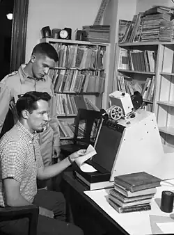 Photographie en noir et blanc de deux personnages concentrés devant un écran, dans une pièce encombrée de documents.