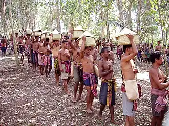Sur le chemin du retour au village. Chaque personne représente un clan. Un clan est composé de plusieurs familles qui, ensemble, forment le village. La main gauche sur la poitrine signifie que la cérémonie est faite de bon cœur, avec honnêteté, confiance et des intentions pures. Tous les hommes ici portent les paniers à la manière des femmes. Dans la vie quotidienne, c'est la façon dont elles transportent leurs marchandises.