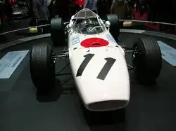 Photographie en couleur de la Honda RA272 blanche avec un cercle rouge, vue de face, dans un musée d'exposition.