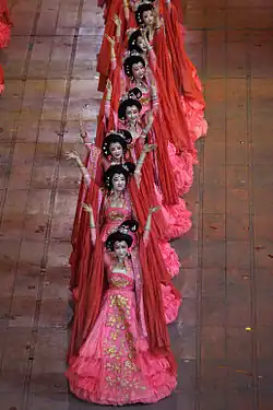 Plusieurs femmes chinoises, dans des vétêments traditionnels, sur une file indienne et les bras en l'air.