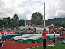 Photo large plan montrant une perchiste en pleine ascension. Des officiels sont au premier plan, la sauteuse se situe au second plan et on aperçoit le public au troisième plan.