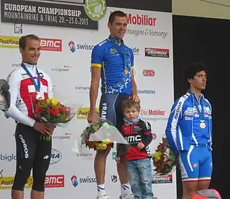  Trois coureurs cyclistes debout sur un podium avec une médaille autour du cou et tenant un bouquet de fleurs.