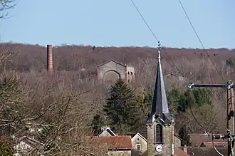 Trois bâtiment en ruines et une cheminée tronquée dépassent des arbres au-dessus de la collines surplombant le village.