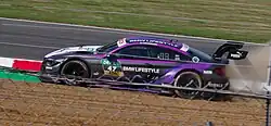 Photographie d'une voiture de tourisme allemande noire et violette, vue de profil gauche, derrière un grillage, projetant quelques graviers derrière elle.