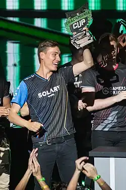 Un joueur de jeu vidéo habillé d'un T-shirt et en jean soulève un trophée sur lequel est écrit TrackMania Cup.