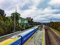 Dans le quart en haut à gauche, un bâtiment de style soviétique, massif et rectangulaire, avec une coupole, domine la scène avec sa façade crème et ses larges fenêtres. Au centre-gauche au premier plan, un train aux wagons bleus et jaunes est à quai, et un autre train sur l'autre côté du quai, rouge, semble rentrer en gare. Le quai en béton s'étire vers l'arrière-plan, où l'on devine des panneaux d'affichage et des lampadaires métalliques, ainsi qu'une passerelle piétonne au centre. Le ciel, pâle et en grande partie nuageux, occupe le tiers supérieur de l'image.