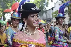 Photographie d'une proccession de rue. Plusieurs drag queens latinas, souriantes, portent des robes colorées, des chapeaux de feutre et des bijoux en perle