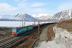 Un convoi de wagons noirs remplis de charbon occupe le centre de l'image, tiré par une locomotive diesel. La voie ferrée, à double voie, longe les rives d'un lac, au centre de l'image, aux eaux gris-bleu qui reflètent un ciel chargé de nuages.  À droite, la berge rocheuse lac est dénudée de végétation haute, seulement des herbes. En arrière-plan, des montagnes haute et pelées, partiellement enneigées, ferment l'horizon.