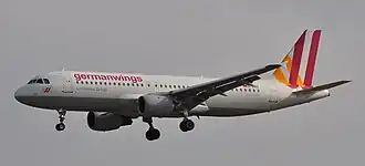 Photo du côté gauche d'un avion de ligne, aux couleurs blanches avec des bandes oranges et rouges sur la queue, en phase d'approche avec le train d'atterrissage sorti.