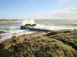 Au premier plan, rochers avec une toute petite jetée. En arrière-plan à gauche, le littoral du continent.