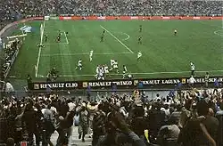 Vue depuis les tribunes du terrain où les joueurs en blancs célèbrent un but devant les supporters.