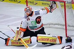Photographie d'un gardien de but en maillot blanc et casque orange arrêtant le palet