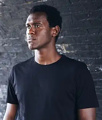 Photo en buste d'un homme noir, portant un haut noir et regardant vers la gauche.