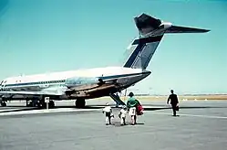 Photographie d'un avion de ligne garé sur le tarmac, avec quatre passagers s'apprêtant à embarquer.