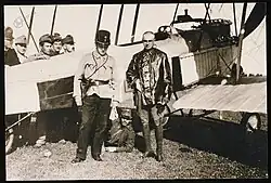 Photo noir et blanc de deux hommes habillés avec un mélange d'uniforme et de tenue de vol devant un avion, avec d'autres militaires derrière l'avion.