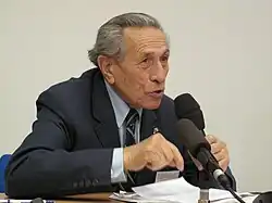 Un homme âgé en costume, assis, un micro face à lui, un autre micro dans sa main gauche.