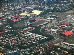 Photographie aérienne d'un complexe sportif comprenant notamment un grand stade de football et plusieurs cours de tennis. L'ensemble est entouré d'habitations rudimentaires.