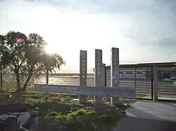 Un monument de béton où est inscrit Justice, Mémoire et Vérité devant un petit hangar à avions et un tarmac.
