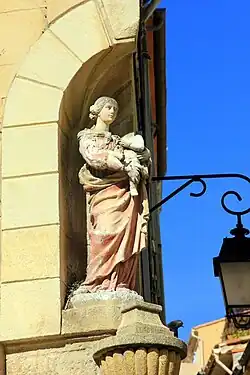 Photographie couleur. Dans une niche en coin, statuette de la Viege portant Jésus qui tient le globe terrestre et une grappe de raisin. La tête de l'Enfant est manquante (avant sa restauration en 2019).