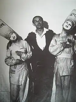 Photographie en noir et blanc. Au centre, un homme de quarante-trois ans, en costume de ville, regarde en l'air et tient par les épaules deux comédiens en costume de scène qui l'encadrent. À gauche, une femme portant un grand bonnet sur lequel est inscrit "reine", à droite un homme portant le même bonnet sur lequel est inscrit "roi". Les deux comédiens, visages blancs, sont très fardés.