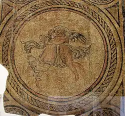 Vue du sujet central de la mosaïque romaine représentant Éros et Psyché : deux très jeunes amoureux ailés sur le point de s'embrasser ; décorations géométriques, dans un vaste rectangle plus haut que large. À l'intérieur des quatre coins, quatre visages inscrits dans des cercles plus petits. Seuls les deux visages de la partie supérieure sont conservés en entier. Un oiseau échassier est représenté marchant vers la gauche au-dessus du cercle central