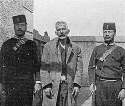Photo en noir et blanc montrant un homme à l'air hagard entouré de deux soldats.