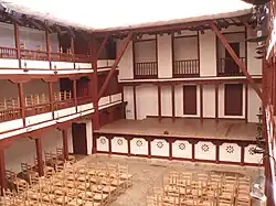 Une cour carrée à ciel ouvert à l'intérieur d'un bâtiment, deux galeries carrées de part et d'autre sont couvertes, au fond une scène surélevée puis un mur avec portes et balcons ; nombreuses chaises.