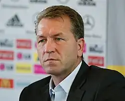 Épaules et visage d'un homme de trois-quart face. Pose en conférence de presse, panneau des sponsors en arrière-plan.