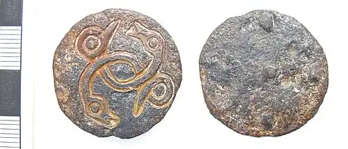 Photo des deux faces d'un disque en métal gris corrodé. Une face est vierge, l'autre porte un dessin gravé représentant deux serpents entrelacés dont le corps fait une boucle avec leur queue en face de leur gueule.