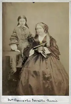 photographie sépia : une femme un peu ronde assise, à gauche de l'image, une petite fille debout