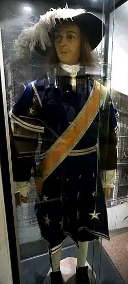 Cliché couleur. Manequin de face en pied représentant un soldt vêtu d'un manteau bleu parsemé depetites étoiles blanches avec une étoile plus importante. Baudrier pour port d'une épéejaune. Chapeau à plume.