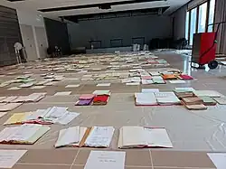 Travail de traitement d'archives suite à une inondation. Les différents feuillets sont étalés afin de favoriser leur séchage. Une bâche en plastique sous les feuillets permet d'éviter qu'ils ne collent au sol. Un extracteur d'humidité facilite l'opération et brasse l'air pour accélérer le séchage, aidé par plusieurs ventilateurs. La salle est fermée pour ne pas apporter d'humidité de l'extérieur. Les archives ont leur côte devant elles afin d'être plus facilement rangées plus tard. Les espacements vides correspondent aux papiers déjà secs et rangés ou à des documents finalement trop abîmés pour pouvoir être conservés.
