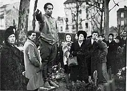 Photographie en noir et blanc d'une foule se recueillant dans un cimetière. Au premier plan, un jeune homme debout sur un rebord pointe son doigt vers le lointain tandis que des gens le regardent.
